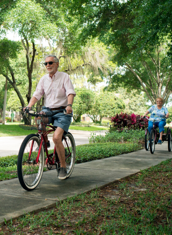 senior man with sunglasses rides bicycle on tree-lined sidewalk, with his wife close behind on a 3-wheel bike