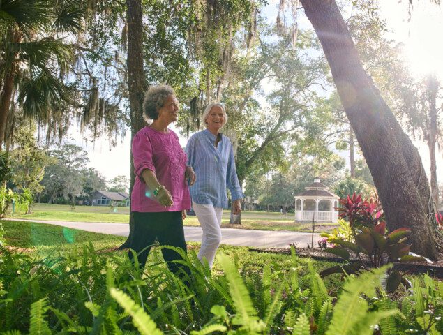 two senior women walk together along a scenic path lined with palm trees and foliage at Village on the Green Senior Living Community